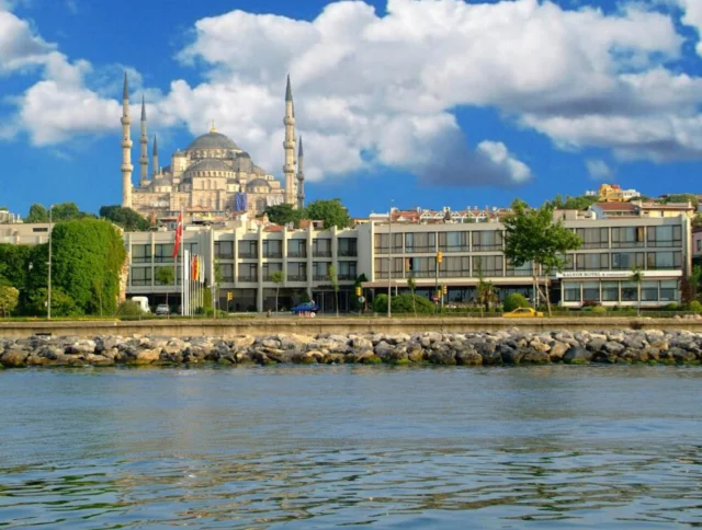 A view of the coastal area with a large mosque featuring multiple minarets in the background under a cloudy sky.