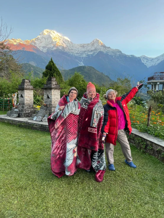 Three people stand on grass in front of snow-capped mountains, two wrapped in blankets. The person on the right points towards the mountains.