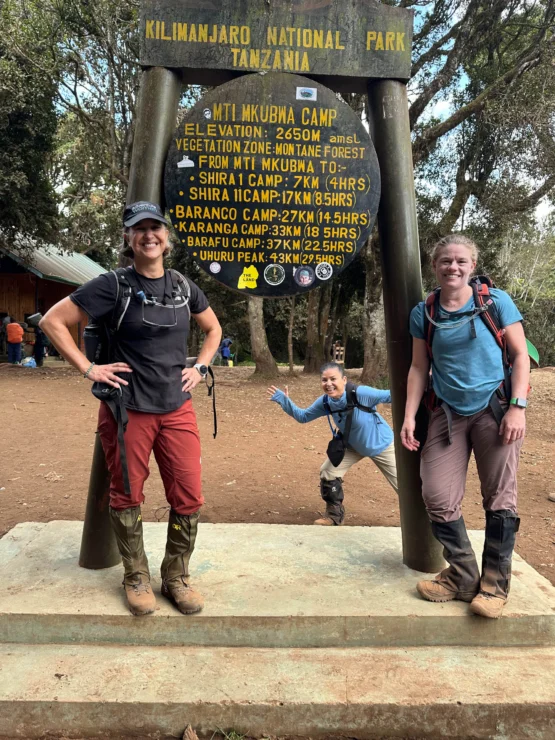 Three people in hiking gear pose in front of a sign at Kilimanjaro National Park, Tanzania. The sign provides elevation details and hiking distances to various camps and peaks.