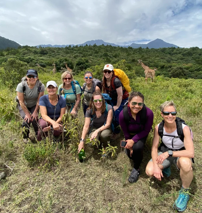 A group of people pose smiling in a grassy field with trees, mountains, and giraffes in the background. They are wearing backpacks and sunglasses.