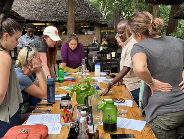 A group of people stand around a wooden table outdoors, examining papers and items. Some are leaning in, appearing engaged in discussion. Trees and a thatched roof are visible in the background.