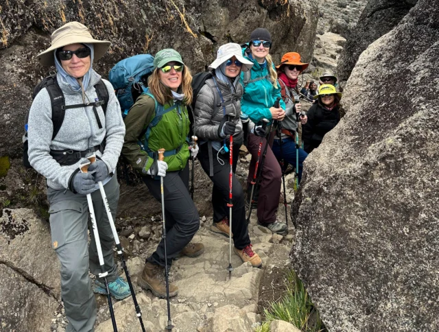 A group of six hikers with trekking poles and gear pose between large rocks on a rocky trail.