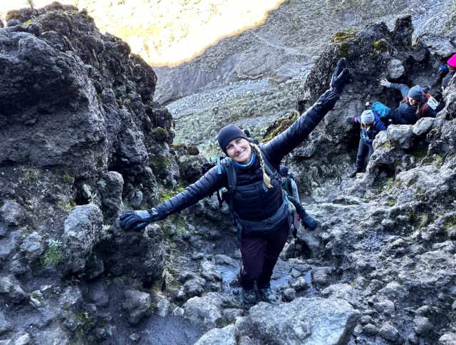 Person in outdoor gear climbing a rocky trail with arms outstretched. Other hikers are visible in the background.