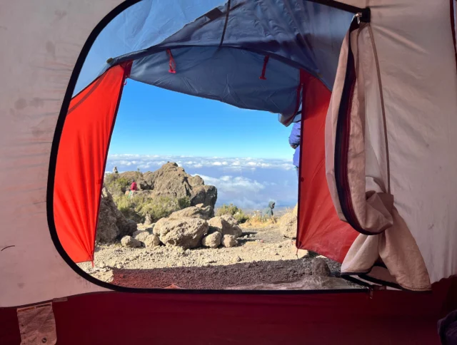 View from inside a tent showing rocky terrain and a clear sky with clouds on the horizon.