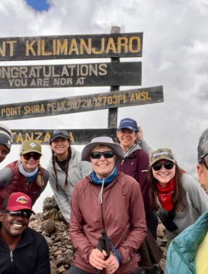 Group of people smiling in front of the Mount Kilimanjaro Shira Peak sign, wearing outdoor gear, under a cloudy sky.
