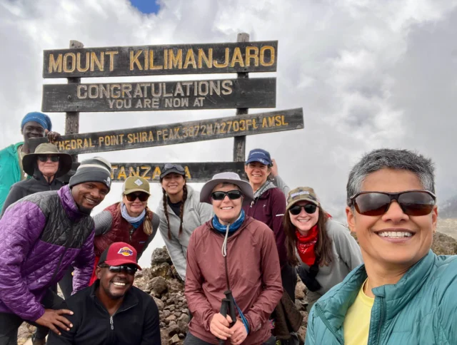 Group of people smiling in front of the Mount Kilimanjaro Shira Peak sign, wearing outdoor gear, under a cloudy sky.