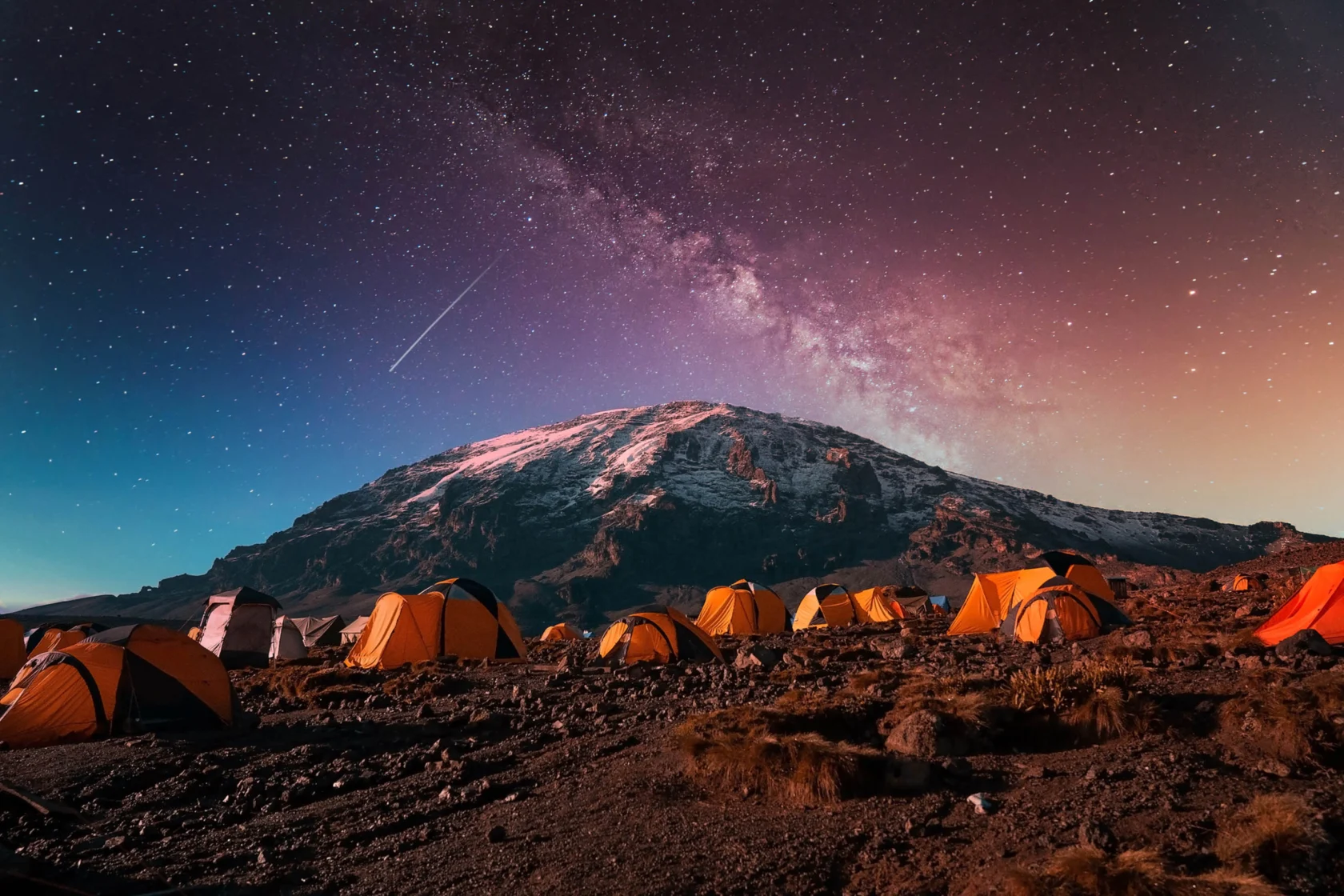 A group of tents on a rocky landscape under a starry night sky with a snow-capped mountain in the background.