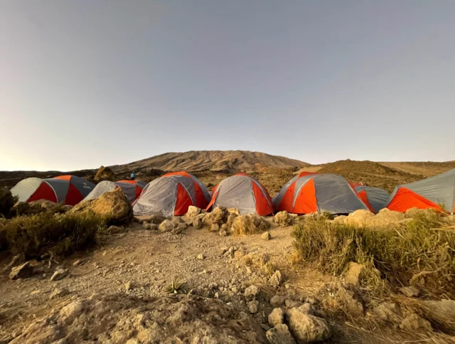 A row of red and grey tents sit on rocky ground with a mountain in the background under a clear sky.
