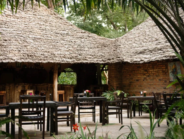 Outdoor dining area with wooden tables and chairs under thatched roofs, surrounded by greenery.
