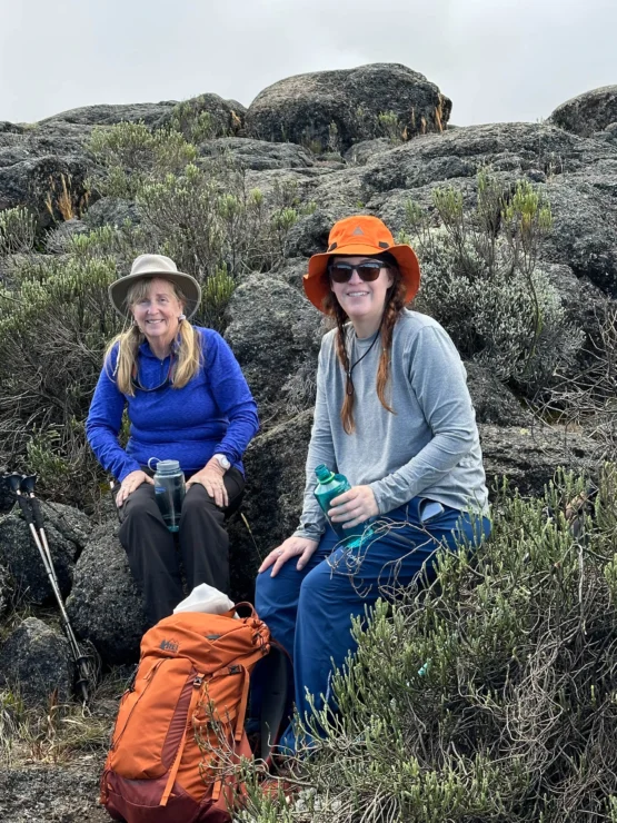 Two people sitting on a rocky terrain, both wearing hats and holding water bottles. An orange backpack is on the ground near them.