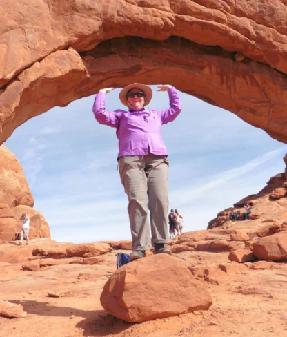 Person in a purple shirt and hat standing on a rock under a natural stone arch in a desert landscape. Other people are visible in the background.