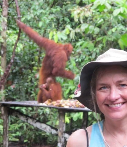 Woman smiling in the foreground with orangutans in the background, set in a lush green forest.