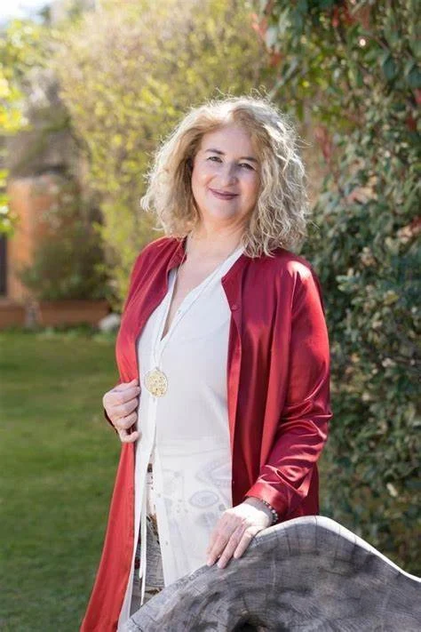 Person with curly hair wearing a red jacket stands outdoors, smiling, with greenery in the background.