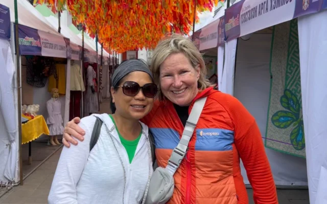 Two people smiling and posing together under a colorful canopy at an outdoor market with tents in the background.