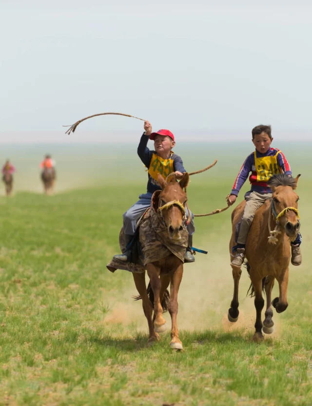 Two people on horseback race across a grassy plain, one raising a whip in the air, with two other riders visible in the distance.