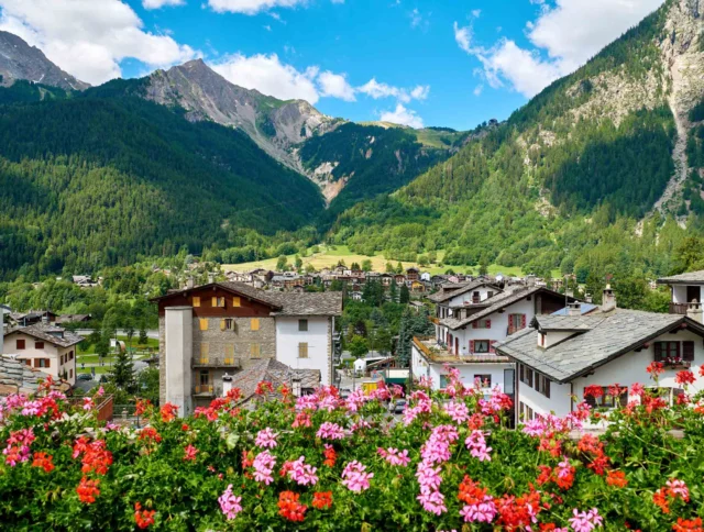 A village with red flowers in the foreground, surrounded by mountains and green forests under a blue sky with clouds.