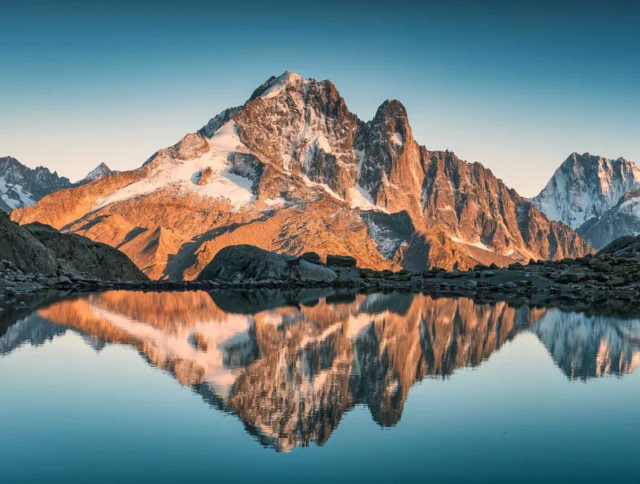 A snow-capped mountain is reflected in a calm lake during sunset, with rocky terrain in the foreground and a clear blue sky above.