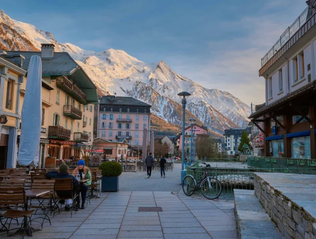 A mountain town scene with people walking along a pathway lined with outdoor cafe tables, bicycles, and buildings, with snow-capped mountains in the background under a clear sky.