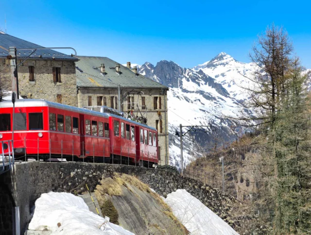 Red train on a mountain track with snow-covered peaks in the background, passing near a stone building.