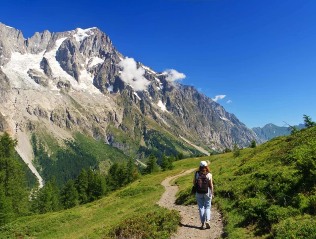 A person with a backpack walks on a trail through a grassy mountain landscape under a clear blue sky.