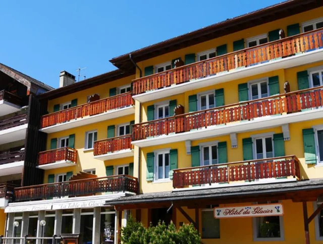 A multi-story yellow hotel with wooden balconies and green shutters under a clear blue sky.