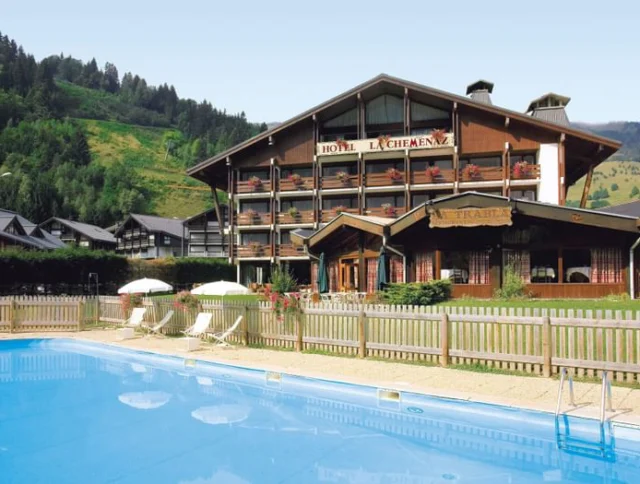 A wooden hotel with balconies in a mountainous area, a fenced pool in the foreground, and green hills in the background.