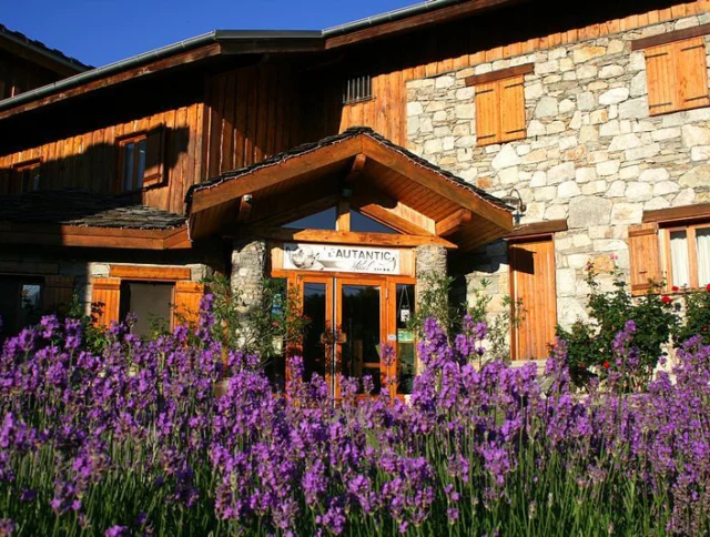 Rustic stone and wood building with wooden shutters, set behind a field of blooming purple lavender under a clear blue sky.