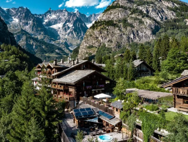 Charming wooden cabins surrounded by lush greenery and mountains under a clear blue sky, with visible solar panels and a small pool in the foreground.