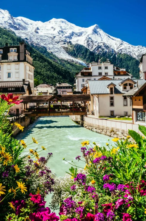 A picturesque alpine village with colorful flowers in the foreground, a river running through the center, and snow-capped mountains in the background.
