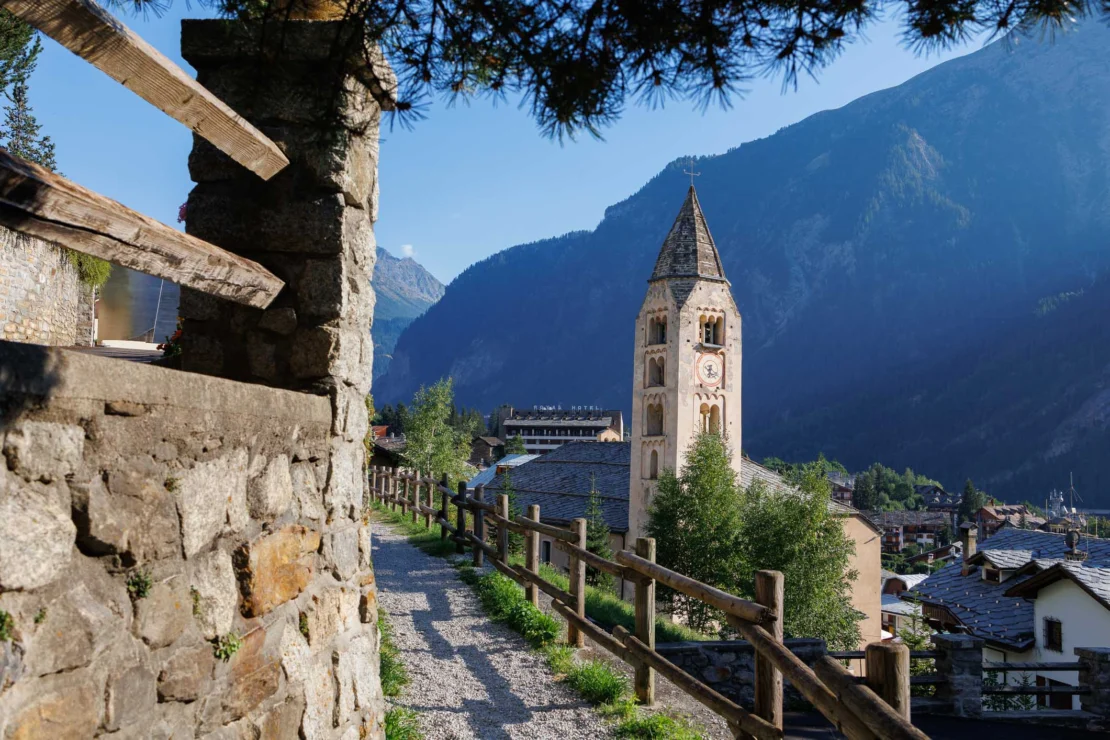 Stone path leading to a church with a tall clock tower, surrounded by mountains and greenery under a clear blue sky.