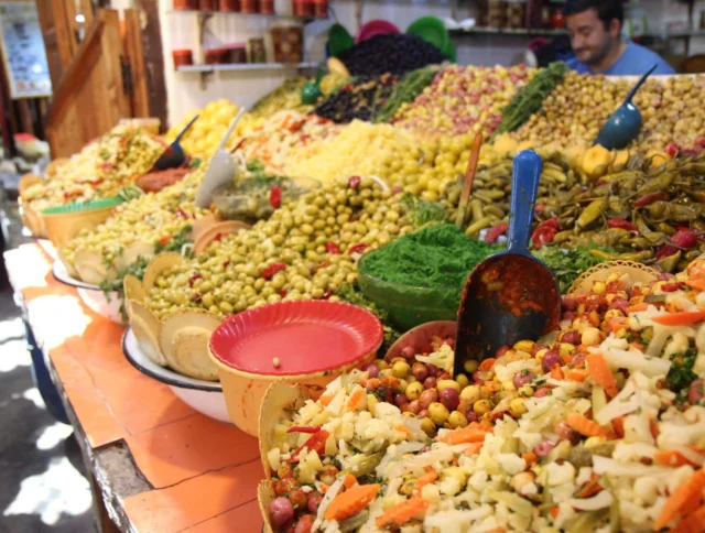 An assortment of pickled vegetables and olives displayed on a market stall, with scoops and bowls, and a vendor in the background.