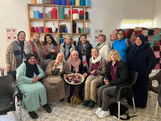 Group of women posing in a room with colorful yarns and threads on shelves, holding handmade items.
