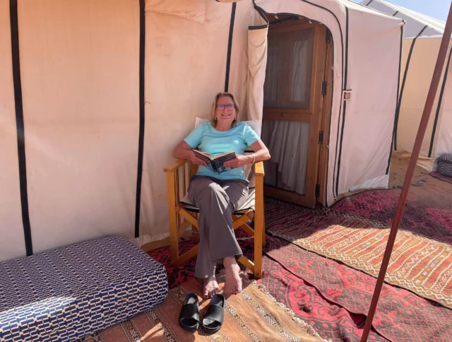 A person sitting in a wooden chair outside a tent, reading a book. Sandals and a cushion are on the ground.
