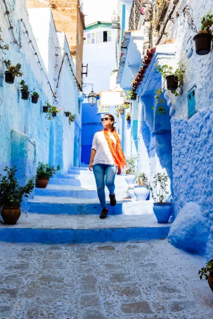Person walking down a narrow blue-painted street with potted plants lining the walls.