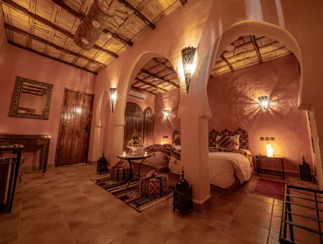 A cozy, warmly-lit bedroom with arched doorways, two beds, decorative lanterns, and a wooden ceiling. The room has tiled flooring and traditional decor.