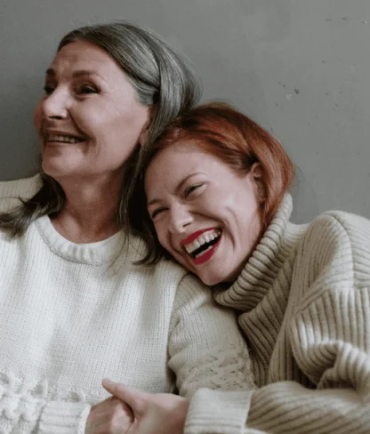 Two women in sweaters, one with gray hair and the other with red hair, smiling and leaning against each other against a plain wall.
