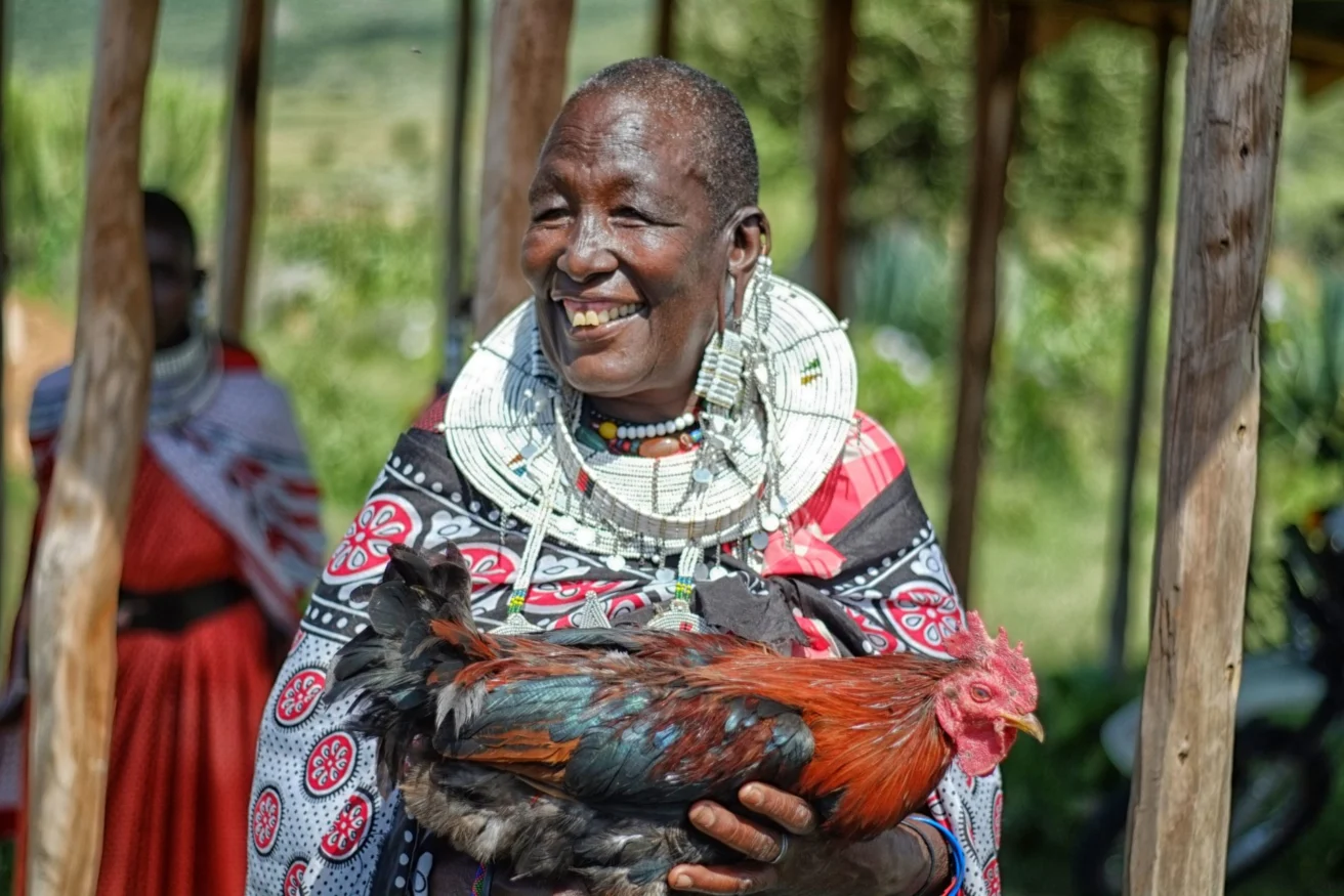 A person wearing traditional attire is smiling and holding a rooster while standing outside near wooden beams.