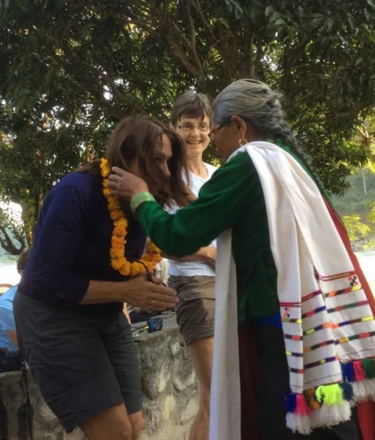 Person in traditional attire puts a flower garland on another person outdoors, with people watching in the background.