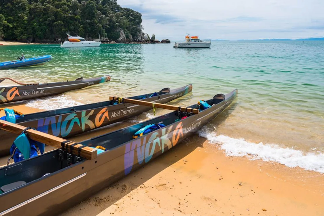 Canoes lined up on a sandy beach with oars, in front of clear turquoise water and a distant boat, under a partly cloudy sky.