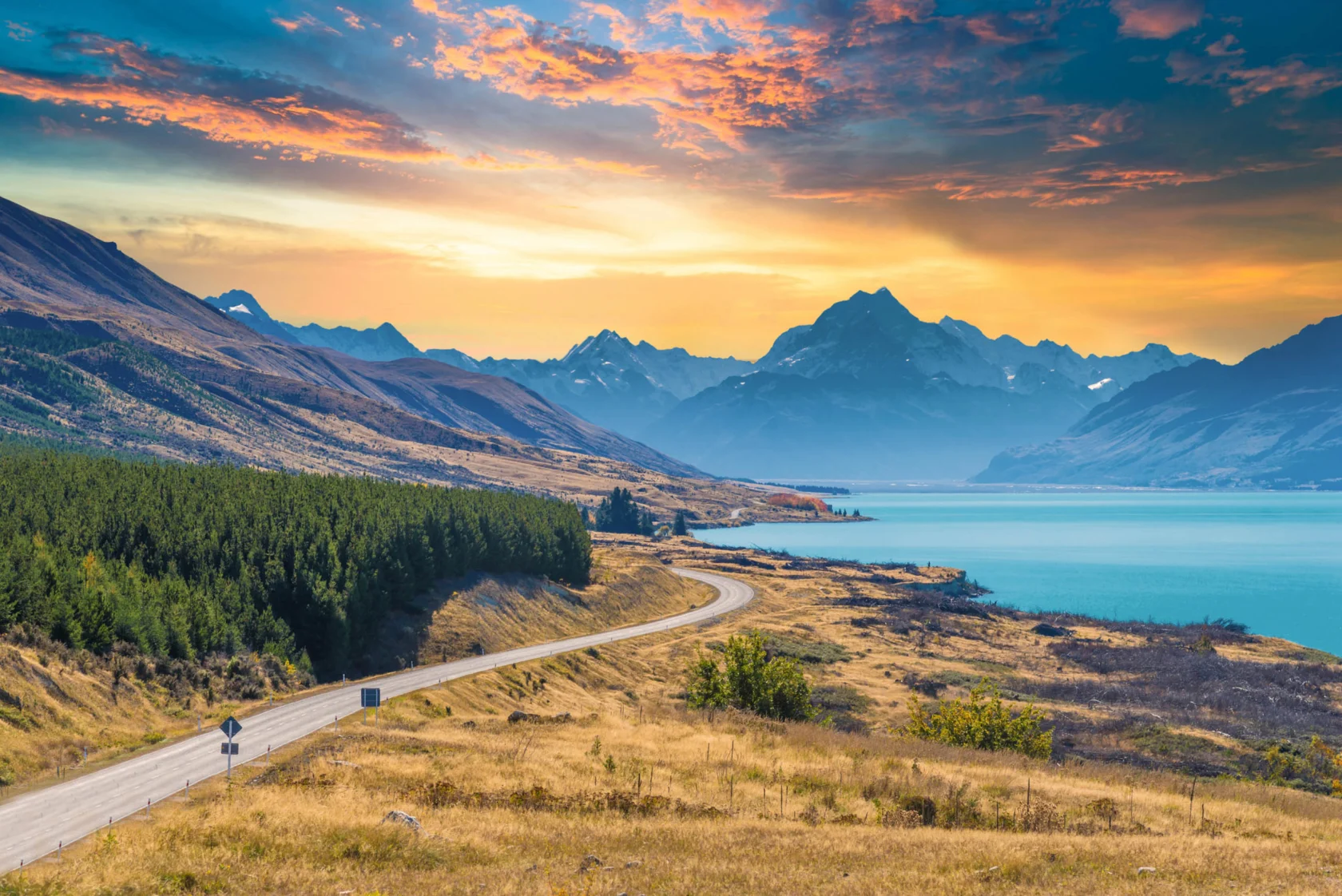 A winding road leads through grassy fields towards mountains at sunset, with a lake on the right and a colorful sky overhead.