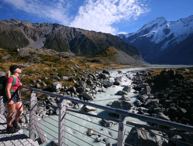 A person stands on a bridge overlooking a rocky river with mountains in the background under a partly cloudy sky.