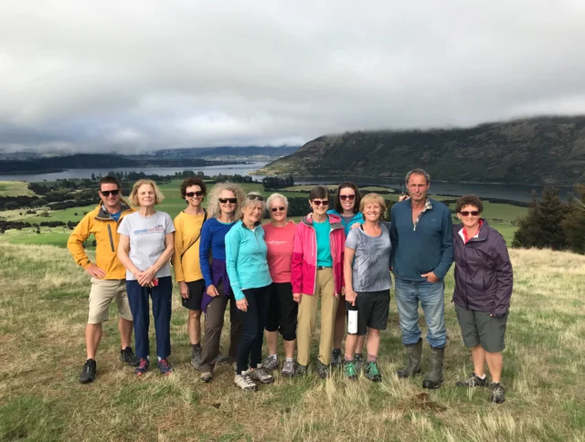 A group of people posing outdoors on a grassy hill with a scenic view of water and mountains in the background, under a cloudy sky.