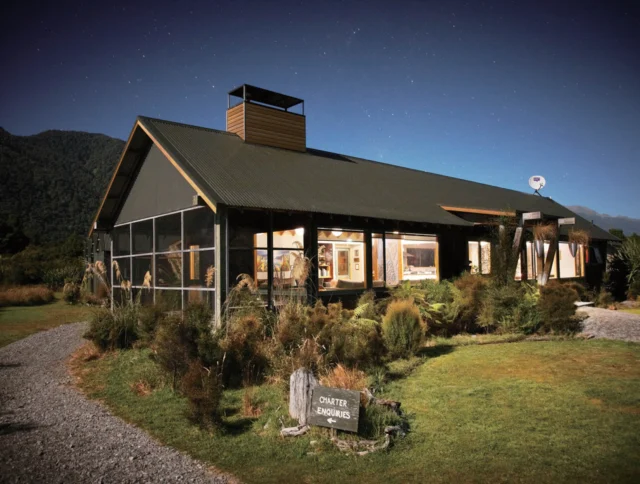 A rustic lodge with a green corrugated roof and large windows is nestled in a wooded area at night. A gravel path leads to the entrance. A sign near the path reads "Cemetery Viewers.