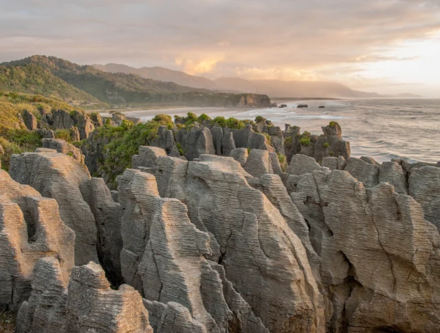 Rock formations at Pancake Rocks, New Zealand, with the ocean and coastline in the background at sunset.