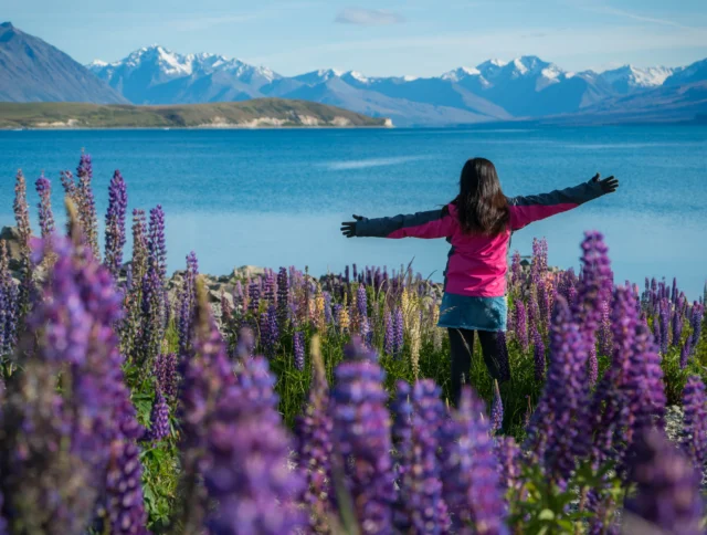 A person stands with arms outstretched in a field of purple flowers by a lake, with snow-capped mountains in the background.