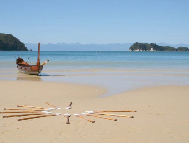 A longtail boat is anchored on a sandy beach. Wooden paddles are laid out in a circular pattern in the sand. Calm sea and islands are visible in the background.