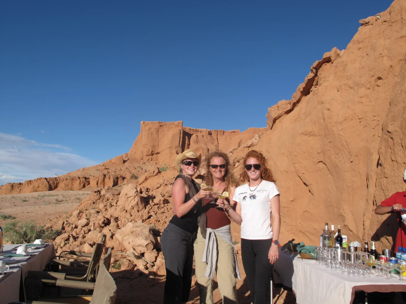 Three people standing outdoors near rock formations, holding drinks and smiling. A table with bottles is nearby under a clear blue sky.