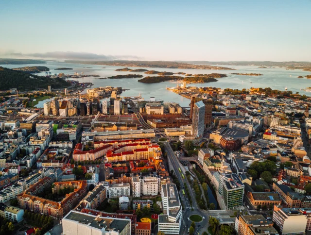 Aerial view of a coastal city with modern and traditional buildings, surrounded by water and greenery, under a clear sky.