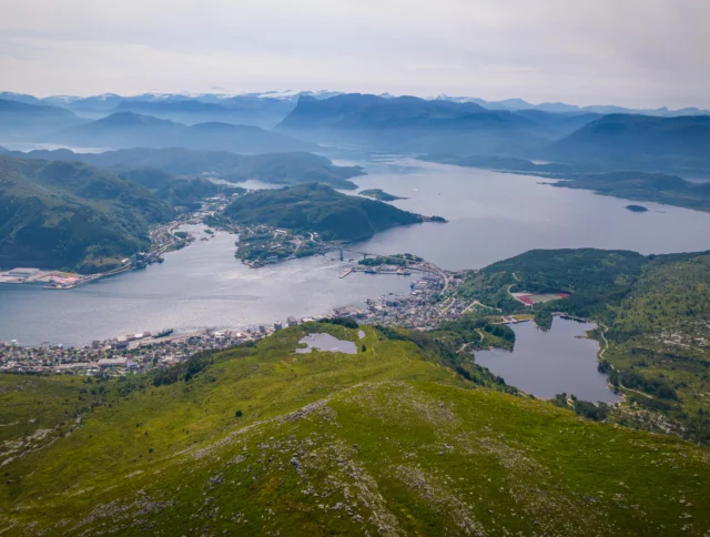 Aerial view of a coastal town surrounded by hills, lakes, and waterways, with distant mountains under a hazy sky.