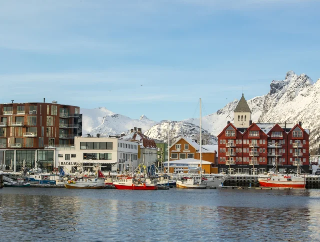 A coastal town with colorful buildings and boats docked along the waterfront, set against a backdrop of snow-capped mountains under a clear sky.